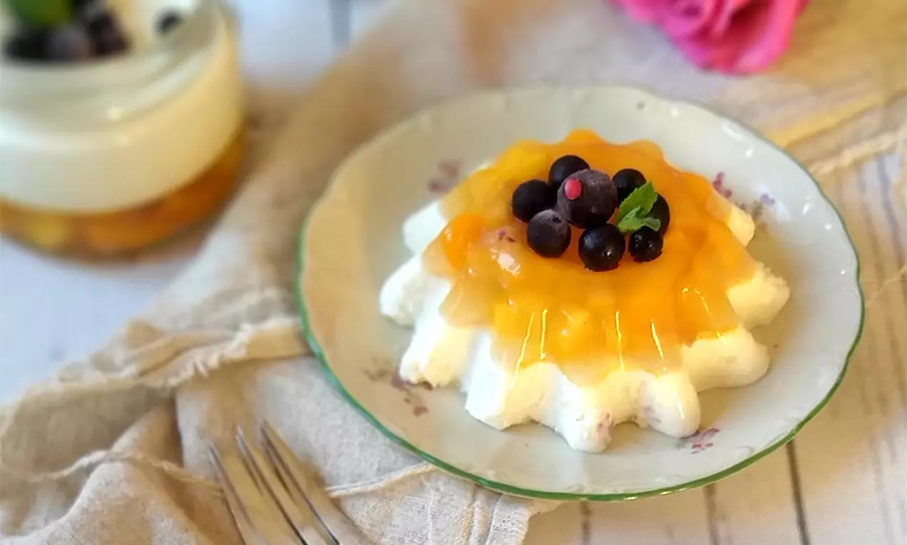Panna cotta zamatovo bielej farby v tvare vlnkovanej kvetiny, pokrytá priezračným mangovým želé s čučoriedkami a lístkom mäty na kvietkovanom tanieri