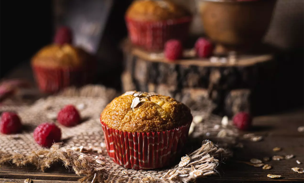 Zlatohnedý muffin v červenom papierovom košíčku na rustikálnej jutovej podložke, ozdobený ovsenými vločkami a obklopený malinami