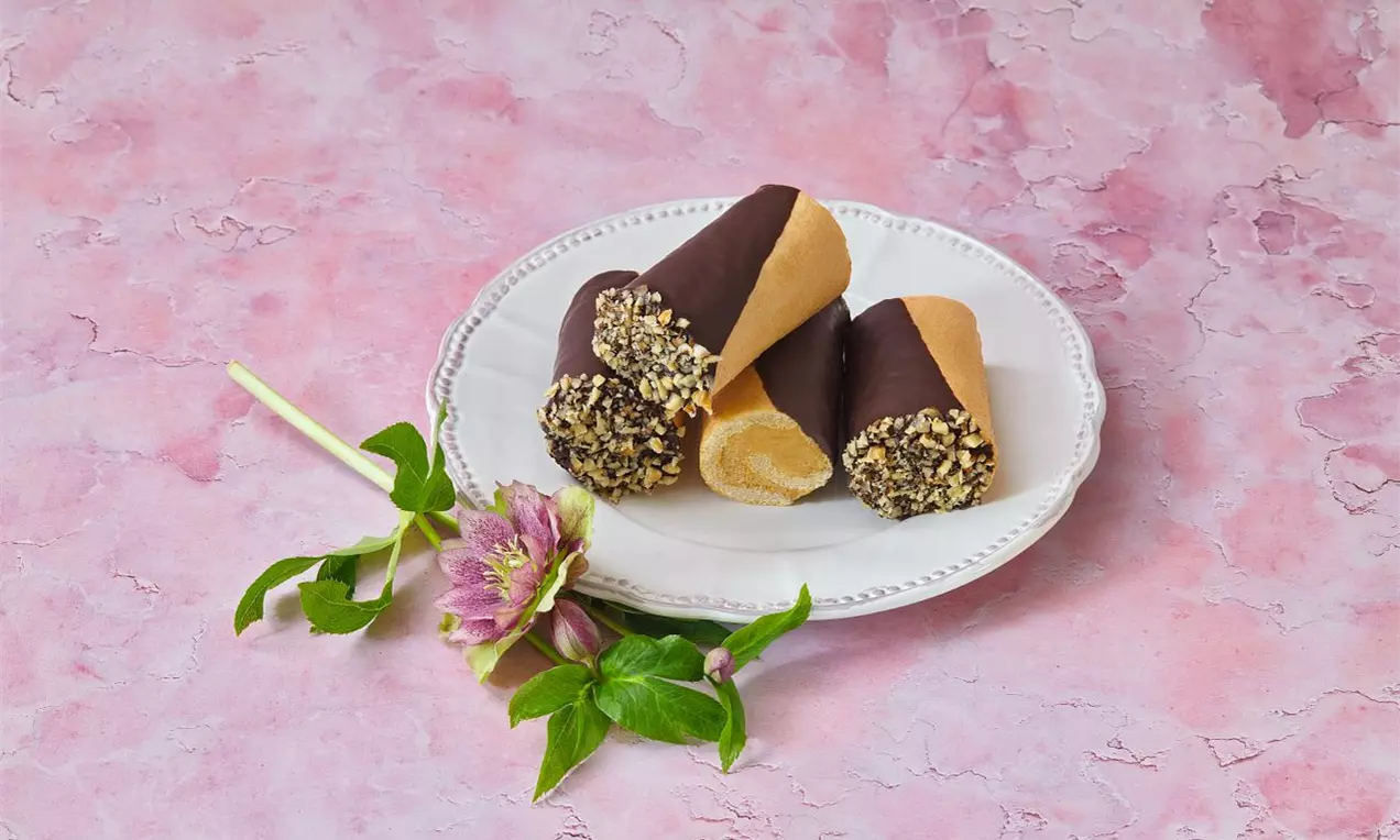 Štafetky zdobené tmavou čokoládou a nasekanými orieškami, na bielom tanieri s fialovým kvetom a zelenými lístkami na ružovom pozadí