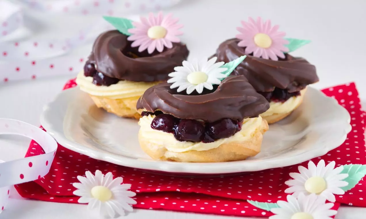 Tri oválne zákusky s čokoládovou polevou a ružovými jedlými kvetmi na bielom tanieri na červenom bodkovanom obrúsku s dekoráciou sedmokrások