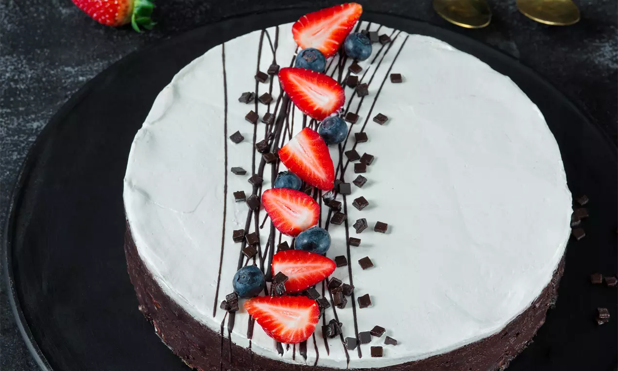 Kakaová torta s tmavým základom, hladkou bielou vrstvou, zdobená čokoládovými hoblinkami, čučoriedkami a jahodovými plátkami na čiernom tanieri