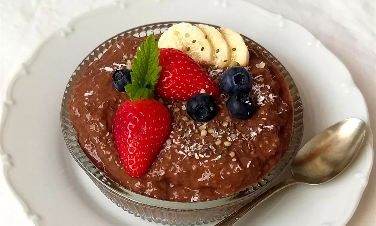 Čokoládový chia puding tmavohnedej farby v sklenenej miske, ozdobený jahodami, čučoriedkami, plátkami banánu, strúhaným kokosom a mätovým lístkom.