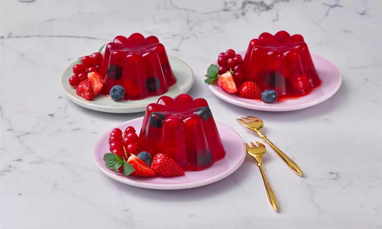 Trojica červených želé báboviek s lesklým povrchom na pastelových tanieroch, doplnená čerstvými jahodami, ríbezľami a čučoriedkam