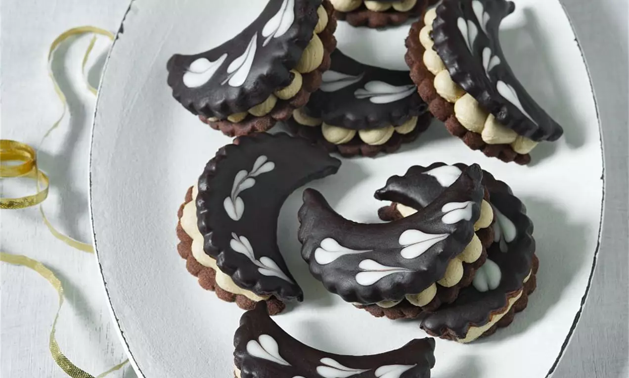 Kávové mesiačiky tmavohnedé s lesklou čokoládovou polevou, bielym vzorom, vrstvené na oválnom bielom tanieri vedľa ozdobnej hviezdy