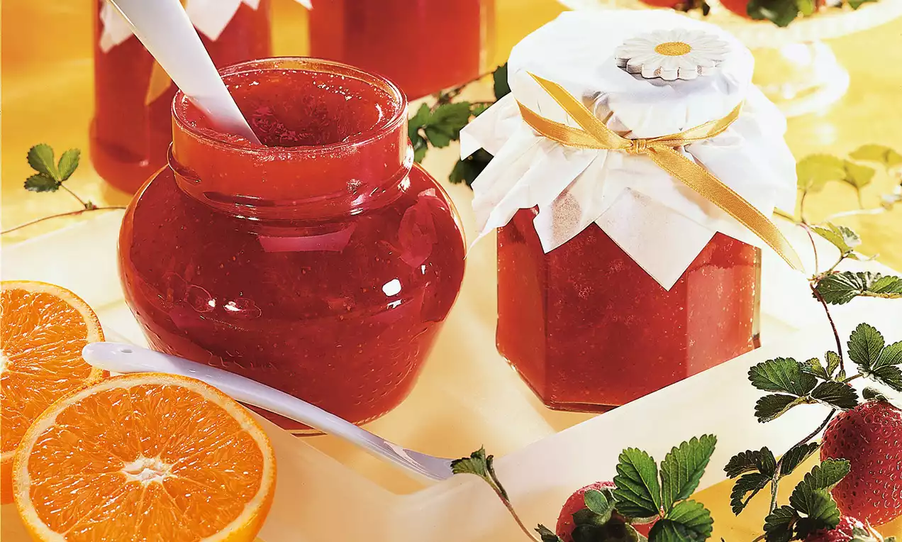 Sklenený pohár s hladkým oranžovo-červeným džemom, vedľa ktorého ležia polovičky pomaranča, jahody a listy na svetlom podnose