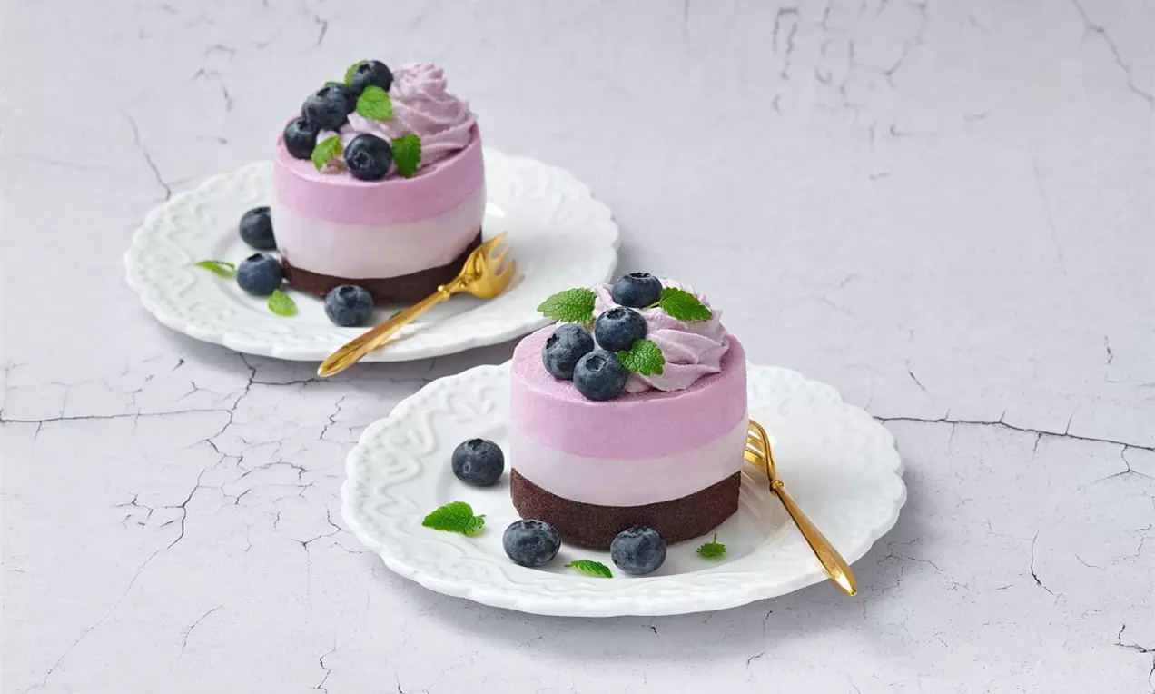 Svetlofialové čučoriedkové tortičky na tmavom sušienkovom základe, ozdobené čerstvými čučoriedkami, lístkami mäty a servírované na bielych tanieroch