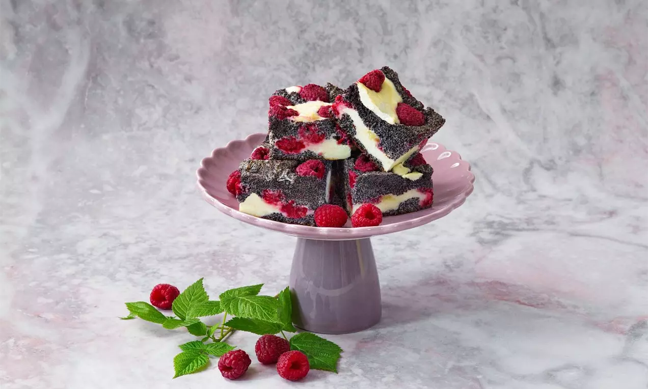 Štvorcové kúsky tmavého makového koláča s vrstvičkami svetlého krému, ozdobené malinami na ružovom podnose, vedľa čerstvej vetvičky malinovníka
