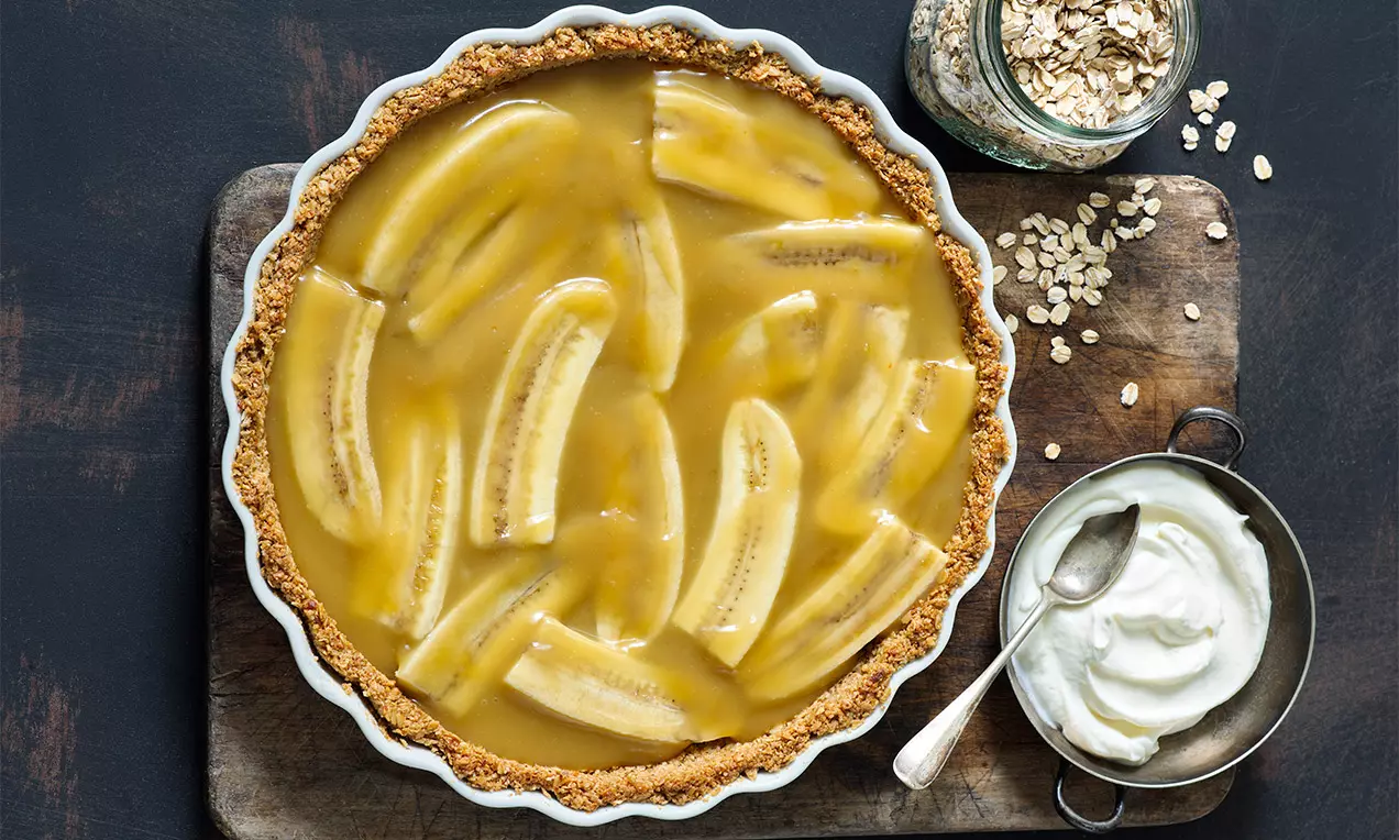 Nízkotučná vegánska banoffee plnka zlatá a hladká, rozpolené banány na chrumkavom hnedom základe v bielej forme na tmavom podklade