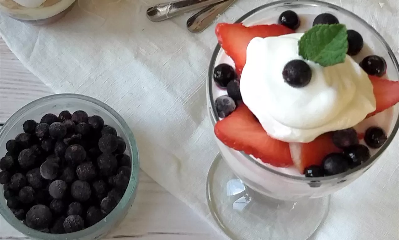 Rybízovo-jahodový trifle vo vysokom priesvitnom pohári, vrstvený krémovo, ozdobený plátkami jahôd, čučoriedkami, šľahačkou a lístkom mäty