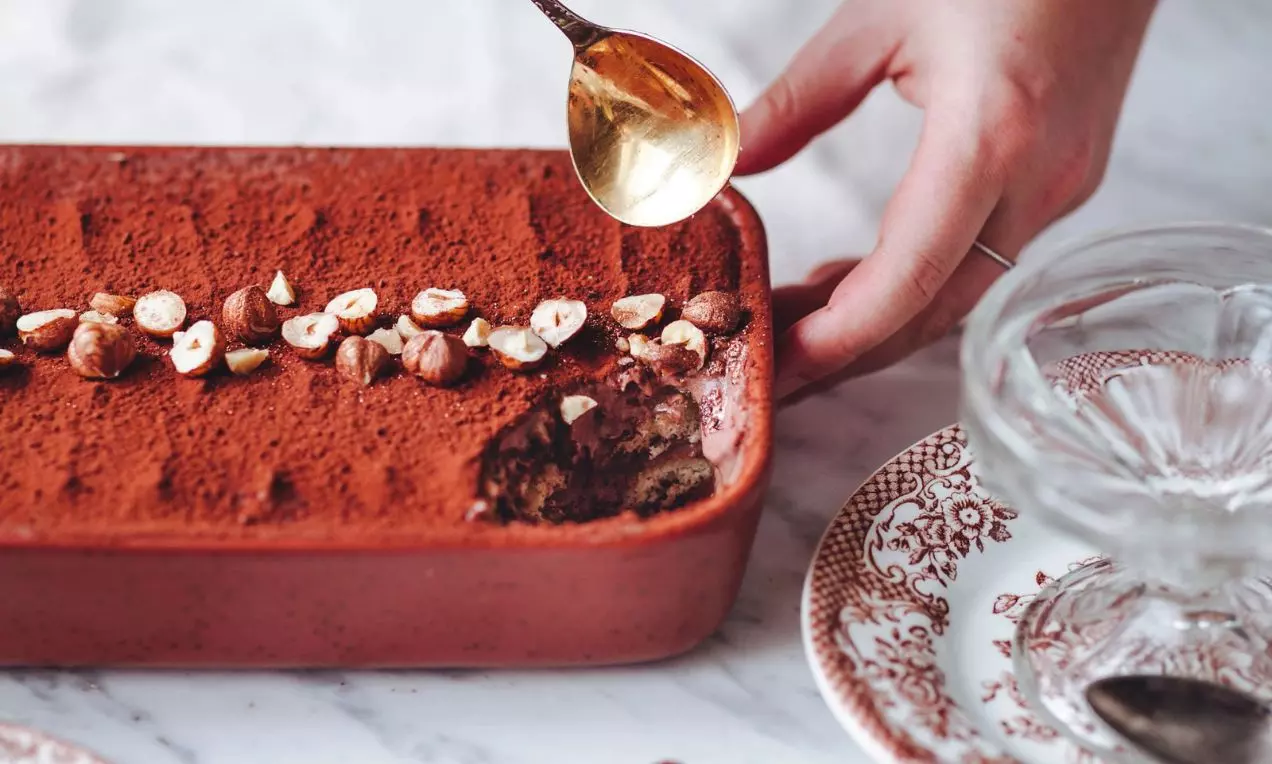 Čokoládovo-lieskoorieškové tiramisu v červenej keramickej forme, posypané kakaom a lieskovými orieškami, s naberačkou a dekorom tanierov na mramorovom stole