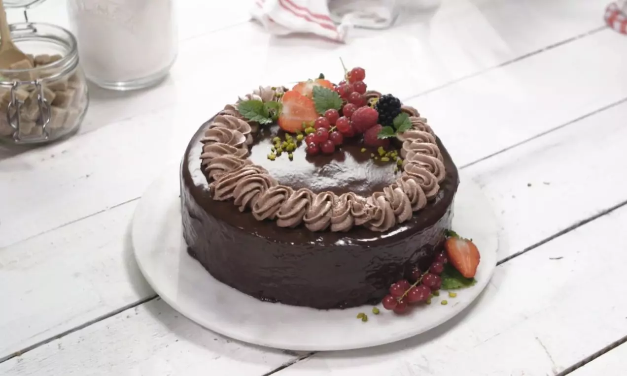 Okrúhla torta s lesklým čokoládovým povrchom, zdobená čokoládovým krémom, čerstvým ovocím a lístkami mäty na bielom porcelánovom podnose