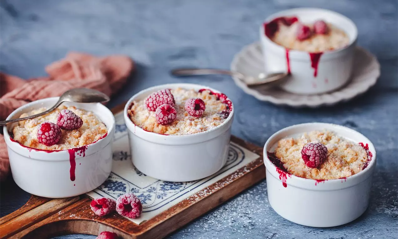 4 mištičky so zapekaným dezertom s posýpkou navrchu, položené na drevenej doske a modrej pracovnej ploche, ozdobené malinami