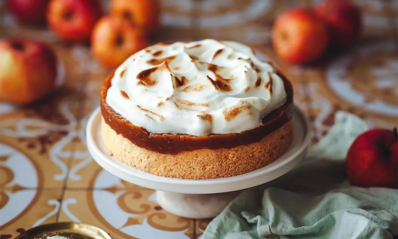 Jablkový koláč so svetlým korpusom, jablčnou vrstvou zdobený snehom a bielym servírovacím tanierom. Okolie zdobené jablkami.