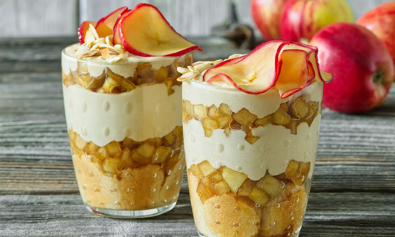 Dva vrstvené poháre z piškótov, jabĺk a krému, zdobené plátkami sušených jabĺk; vo vysokej sklenici na drevenej doske s čerstvými jablkami v pozadí.