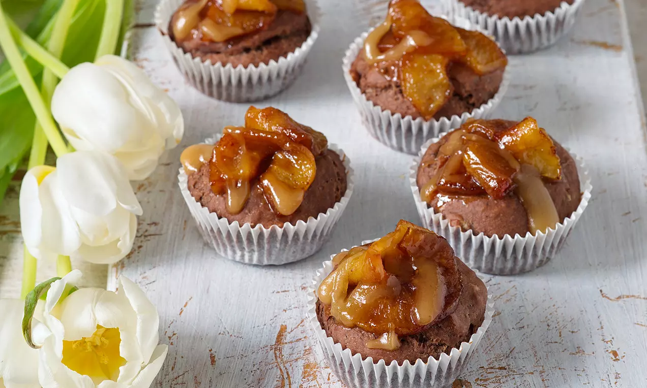 Hnedé čokoládové muffiny v bielych košíčkoch, ozdobené karamelizovanými zlatistými plátkami jabĺk, usporiadané na rustikálnom drevenom povrchu