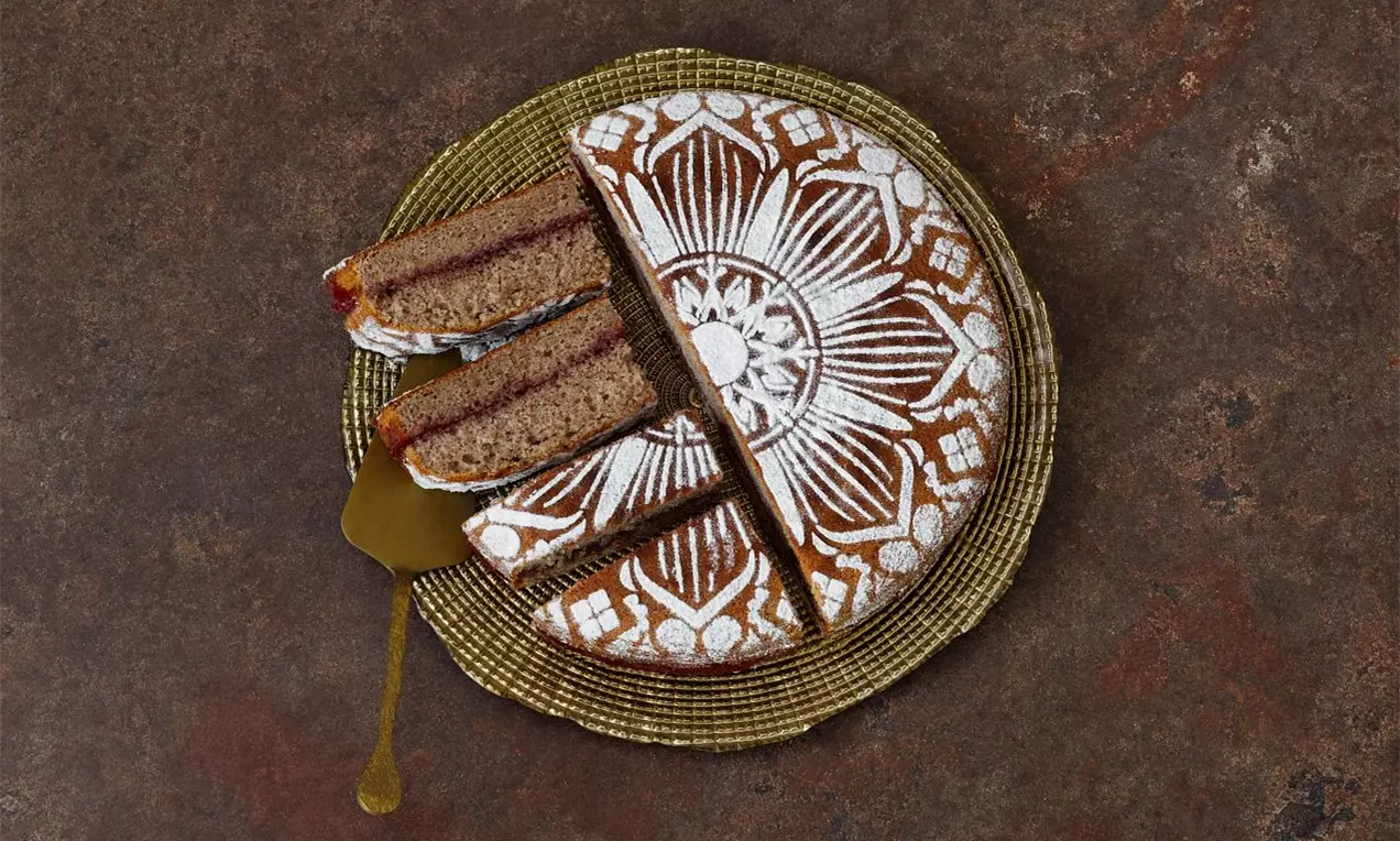 Pohánkový koláč okrúhleho tvaru s výrazným bielym vzorom pripomínajúcim kvetinu, s niekoľkými tortovými kúskami na zlatom podnose