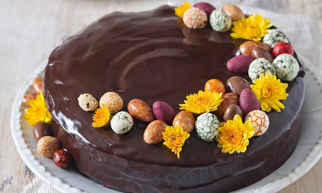 Tmavohnedá čokoládová torta s hladkým lesklým povrchom, ozdobená farebnými vajíčkami a žltými kvetmi na bielom tanieri