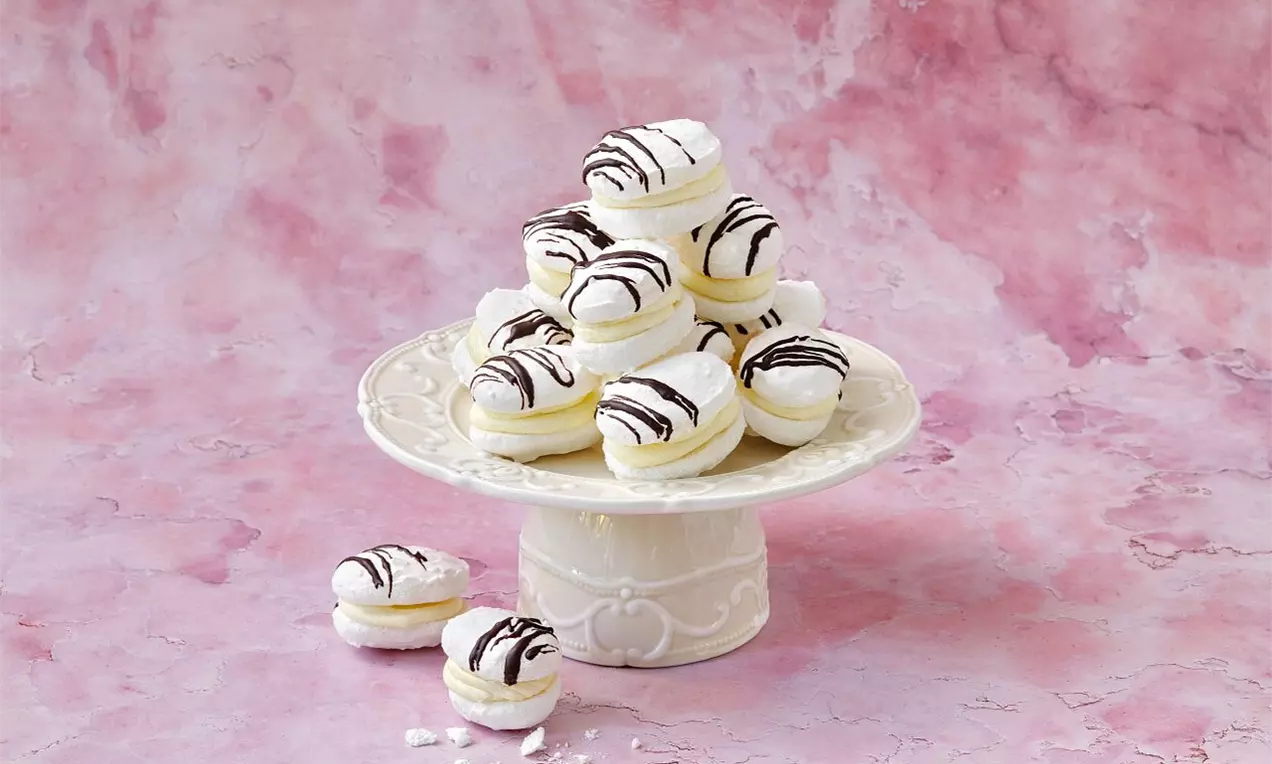 Na svetlom podstavci sú vrstvené biele laskonky oválneho tvaru s kokosovým povrchom, plnené krémom a zdobené prúžkami čokolády
