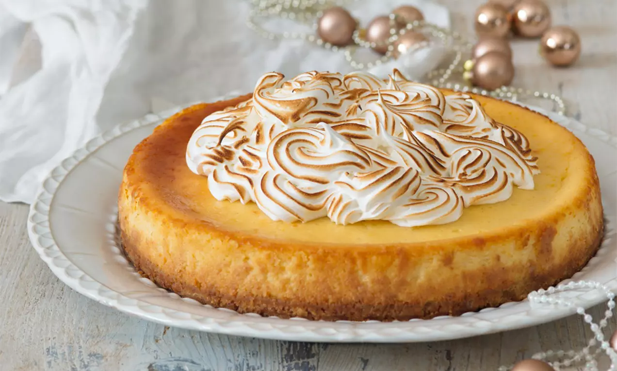Citrónový cheesecake s hladkým žltkastým povrchom, ozdobený dozlatista opraženými pusinkami na bielom vrúbkovanom tanieri
