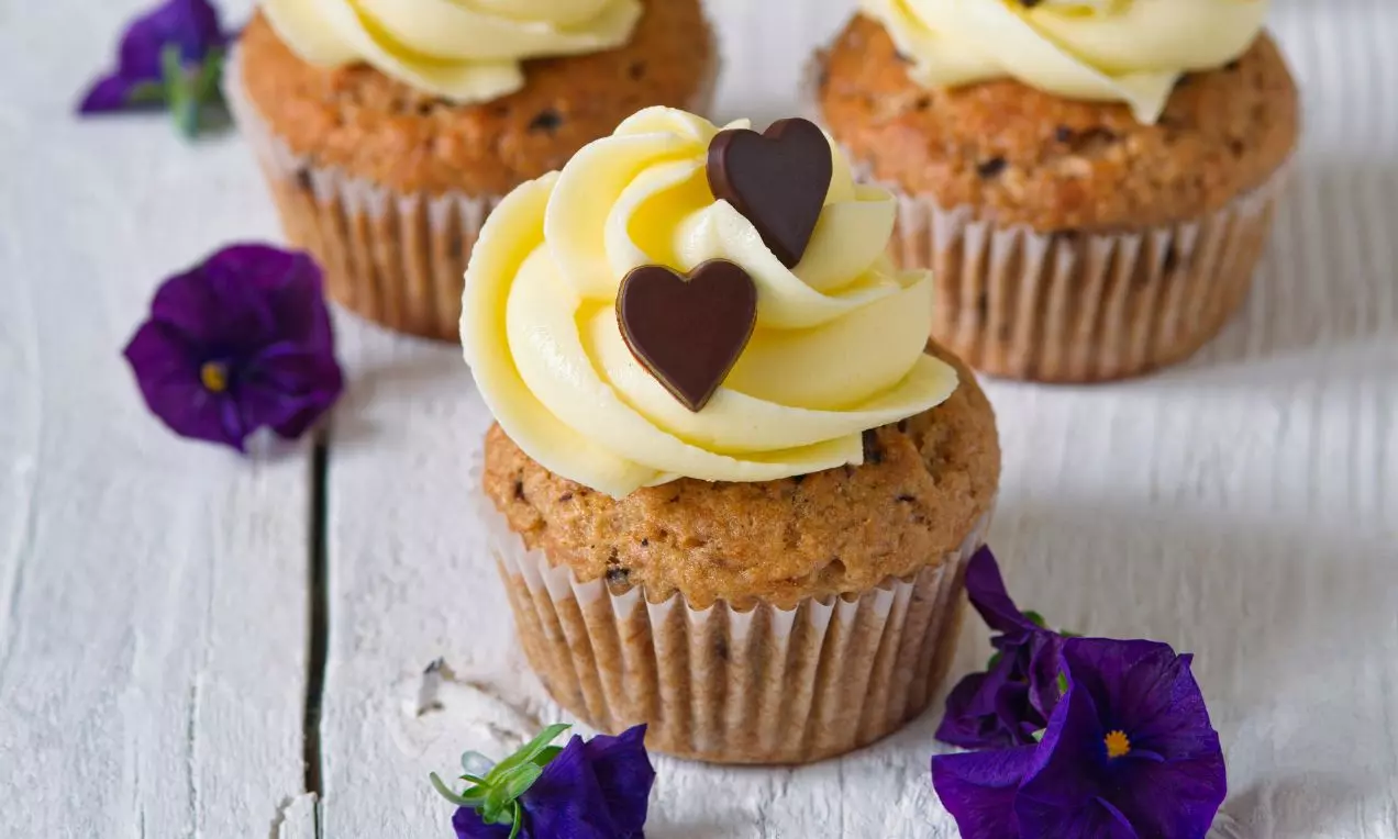 Banánové cupcakes s jemným krémom bledosivej farby, ozdobené čokoládovými srdiečkami, položené na svetlom drevenom podklade vedľa fialových kvetov