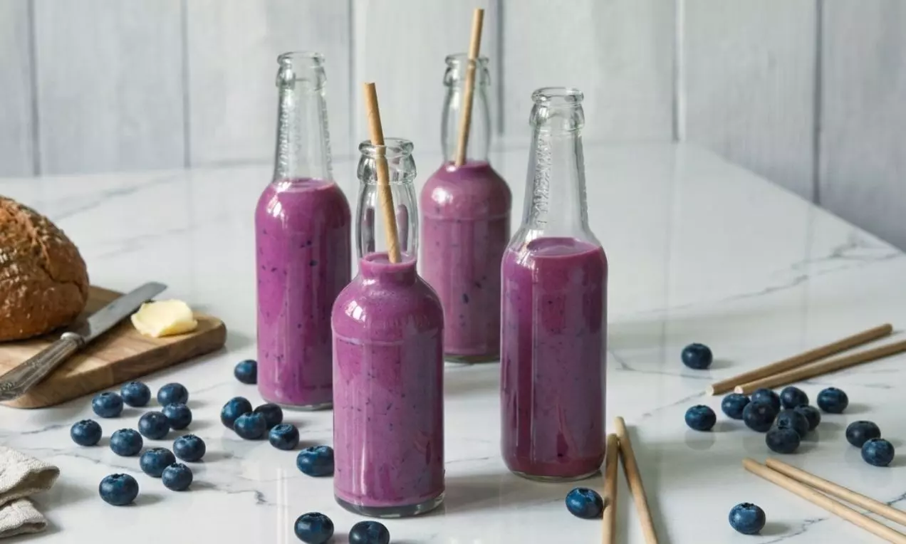 Proteínové smoothie sýtej fialovej farby v sklenených fľašiach so slamkami, obklopené čučoriedkami na bielom mramorovom stole