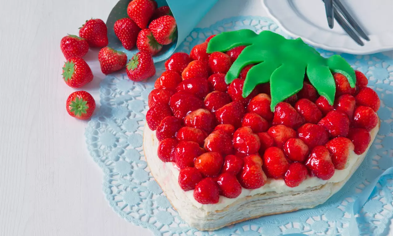 Torta v tvare obrej jahody pokrytá lesklými červenými jahodami a zelenými marcipánovými lístkami na krajkovom obruse