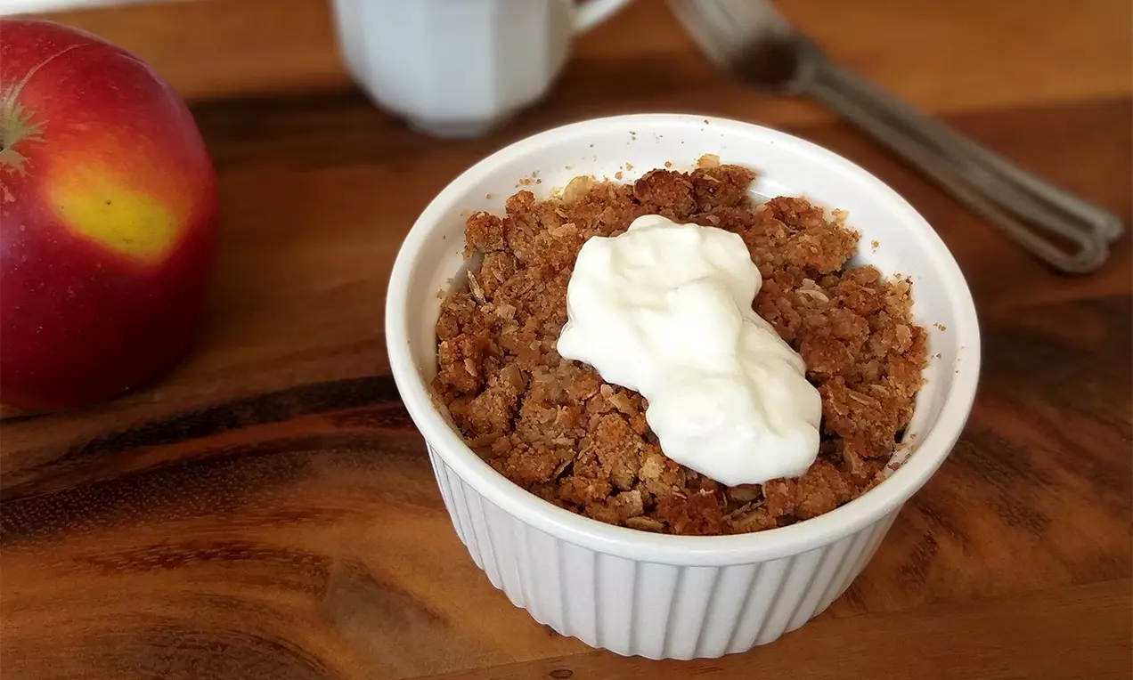 Hnedastý jablečný crumble s chrumkavou drobenkou, ozdobený kopčekom bieleho krému v bielej miske na drevenom stole vedľa červenej jablka