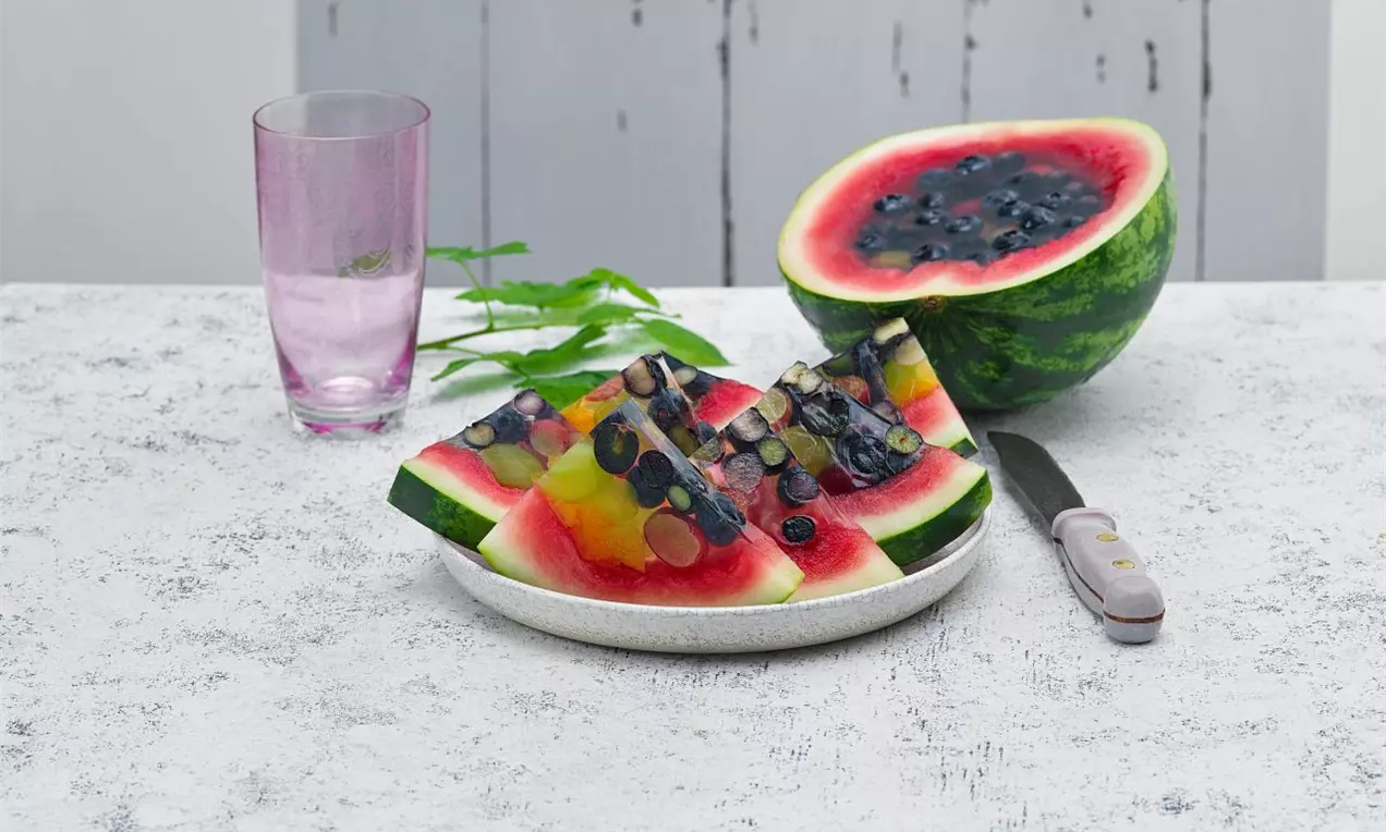 Trojuholníkové kúsky melónu plnené farebným vrstveným ovocným želé s čučoriedkami a plátkami ovocia na bielom tanieri, vedľa pohára vody