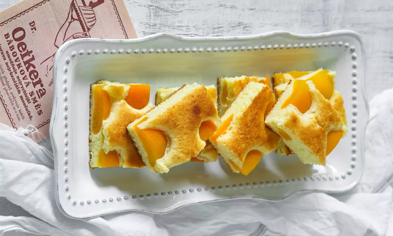 Kúsky meruňkového koláča so zlatistou, nadýchanou piškótou a šťavnatými oranžovými polovicami meruniek na bielom porcelánovom podnose