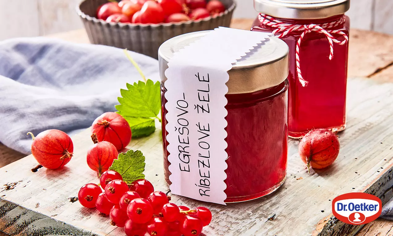 Sýtočervené egrešovo-ríbezľové želé v hladkom sklenenom pohári so zlatým viečkom, obklopené čerstvými bobuľami egreša a ríbezlí na drevenom stole