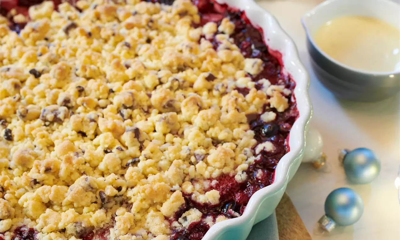Vianočný crumble s rubínovo červeným ovocným základom a zlatistou mrveničkou, uložený v vrúbkovanom bielom tanieri, doplnený miskou krému