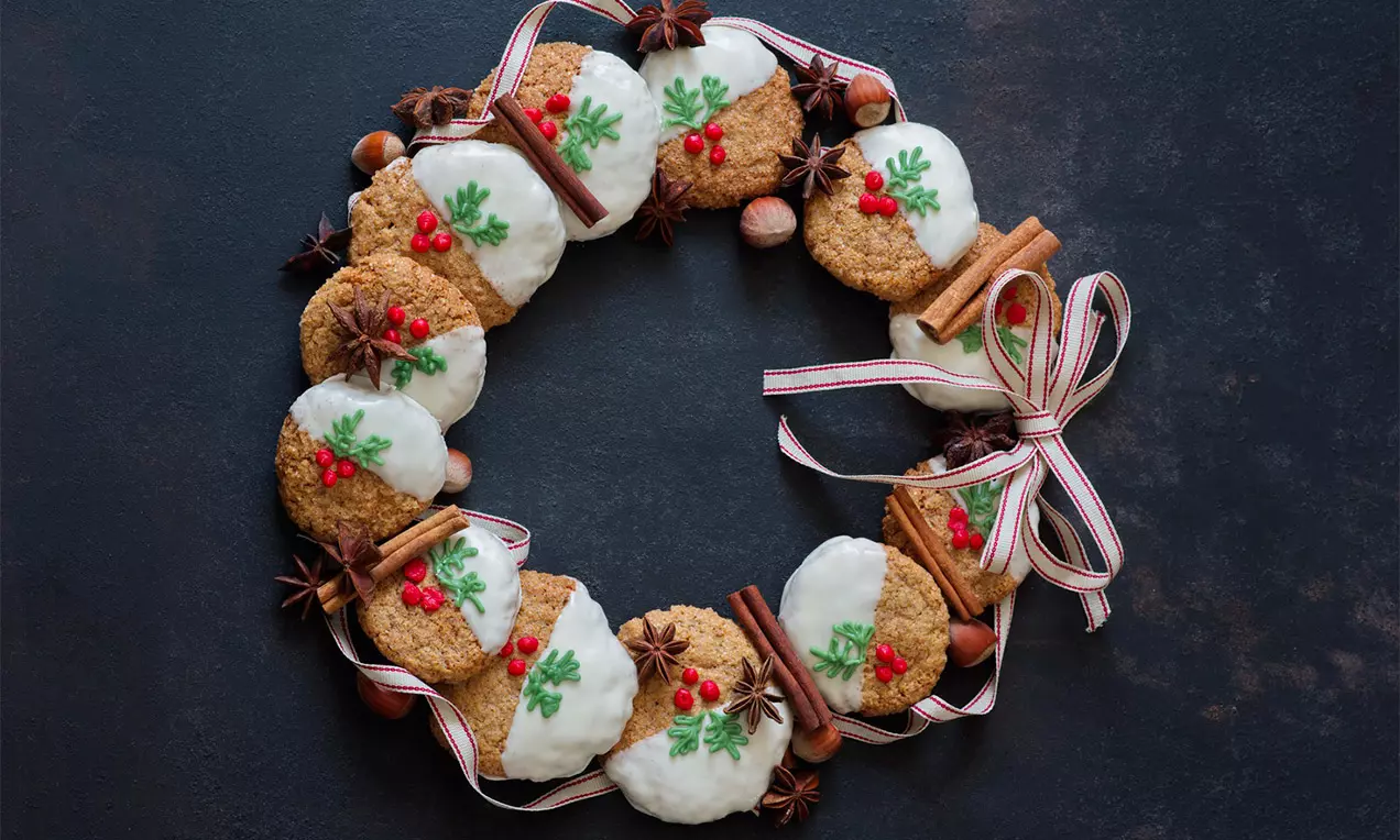 Perníkové cookies hnedastej farby, s bielou polevou, zdobené zelenými lístkami a červenými bobuľami, usporiadané do venčeka na tmavom podklade